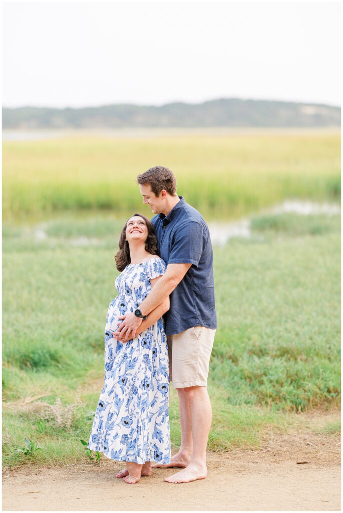 A couple embraces at the Great Island Trail in Wellfleet, MA. The pregnant woman, wearing a blue and white floral dress, smiles up at her partner while he gently holds her belly—a perfect moment for a Cape Cod maternity photographer to capture.