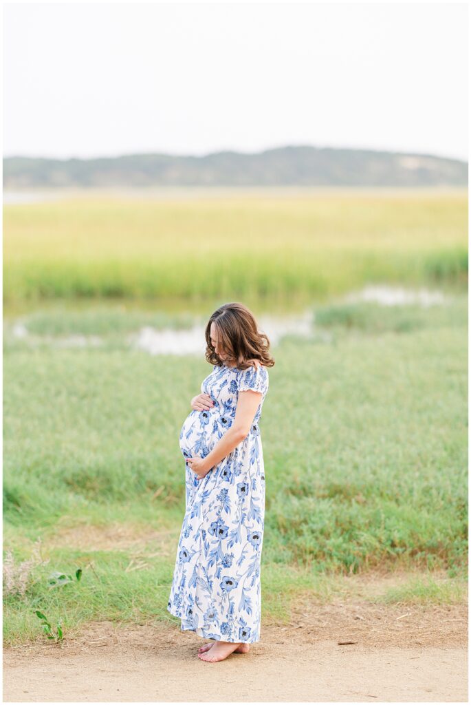 A pregnant woman in a blue and white floral dress stands barefoot on a sandy path at the Great Island Trail, cradling her belly as she looks down lovingly. The grassy marshland in the background creates a peaceful, natural setting.