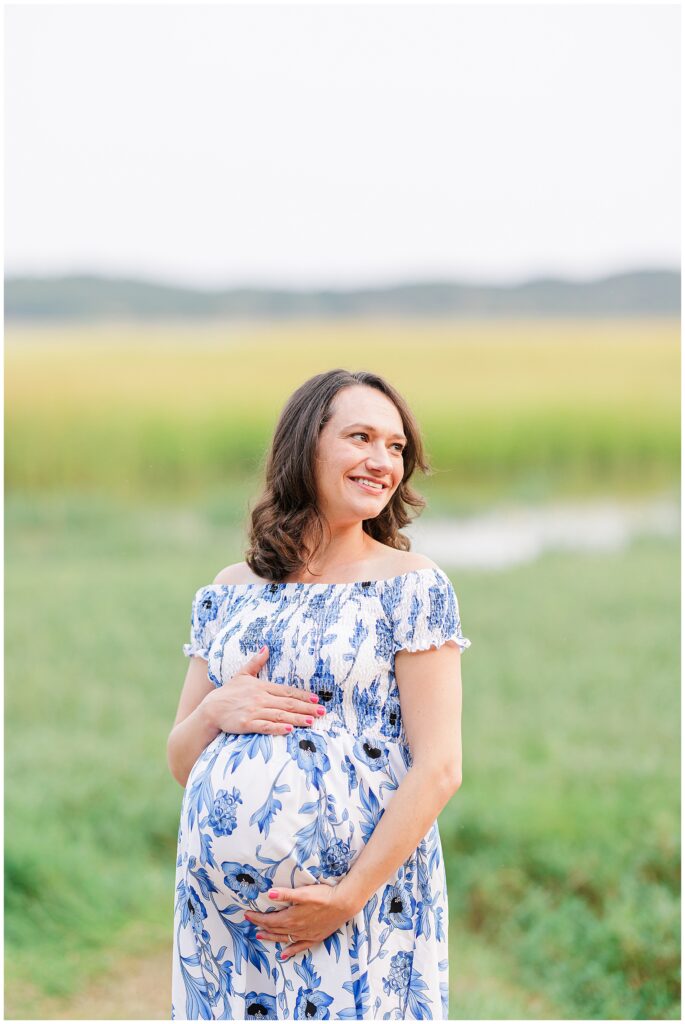 A smiling pregnant woman stands in front of a marsh at Great Island Trail, cradling her belly. Her off-the-shoulder, blue floral dress complements the serene environment, ideal for Cape Cod maternity photography.