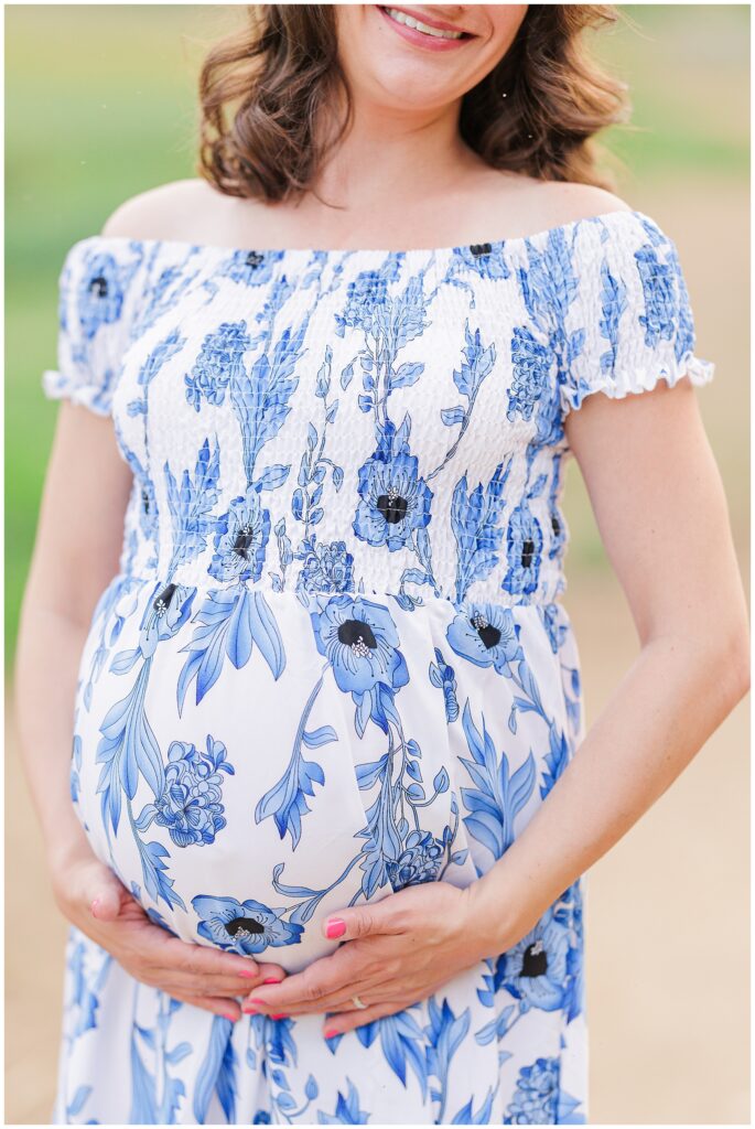 Close-up of a pregnant woman’s belly, lovingly cradled by her hands, in a blue and white floral dress. The background fades softly, highlighting this intimate maternity moment at Great Island Trail.