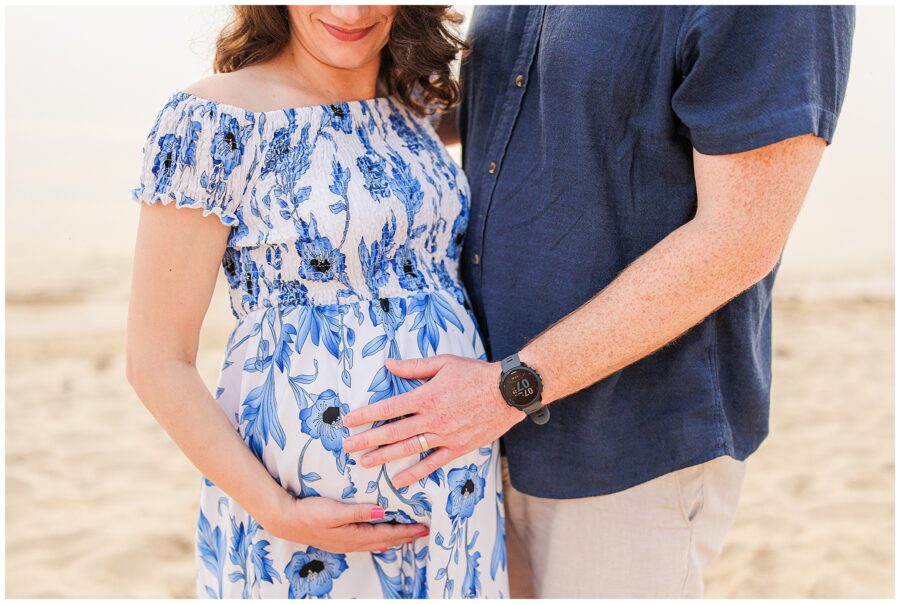 A close-up of a pregnant woman’s belly, adorned in a blue and white floral dress. Her partner’s hand rests on her stomach as they stand on the beach, a beautiful moment captured by a Cape Cod maternity photographer.