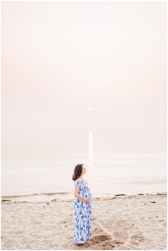 A pregnant woman stands alone on the beach at sunset, gently holding her belly. The soft pastel sky and reflection on the water create a calming backdrop for Cape Cod maternity photography.