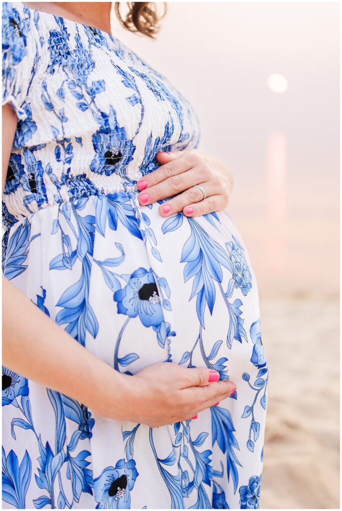 Close-up of a pregnant woman cradling her belly in a blue and white floral dress. Her hands gently rest on her stomach, with the setting sun casting a warm glow in the background at Great Island Trail.