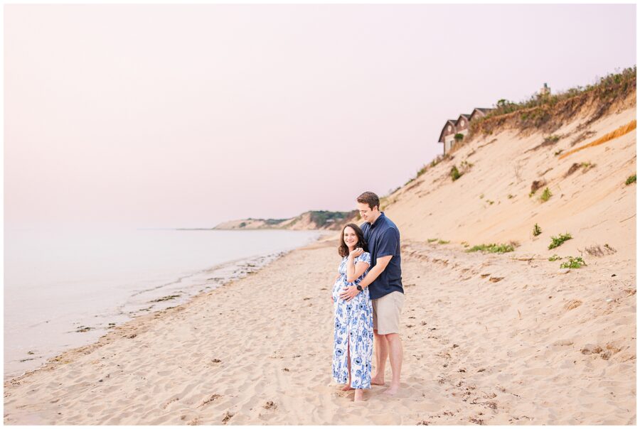 A couple stands together on the sandy shores of Great Island Trail in Wellfleet, MA. The expecting mother, in a blue and white floral dress, is embraced by her partner as they enjoy the calm beach at sunset—an ideal scene for a Cape Cod maternity photographer.