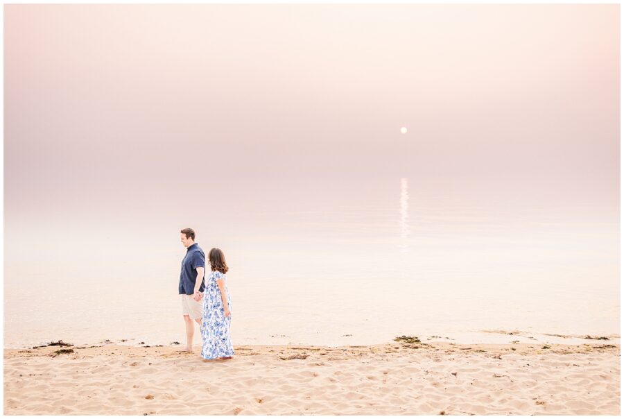 The couple walks hand in hand along the peaceful beach at Great Island Trail, with the soft light of the setting sun reflecting on the water. This tranquil moment highlights the beauty of Cape Cod maternity photography.