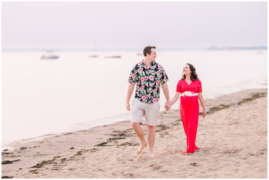 The couple walks barefoot along the shoreline, laughing and holding hands. The expecting mother wears a bright red dress with floral accents, creating a vibrant scene for a Cape Cod maternity photographer at Great Island Trail.