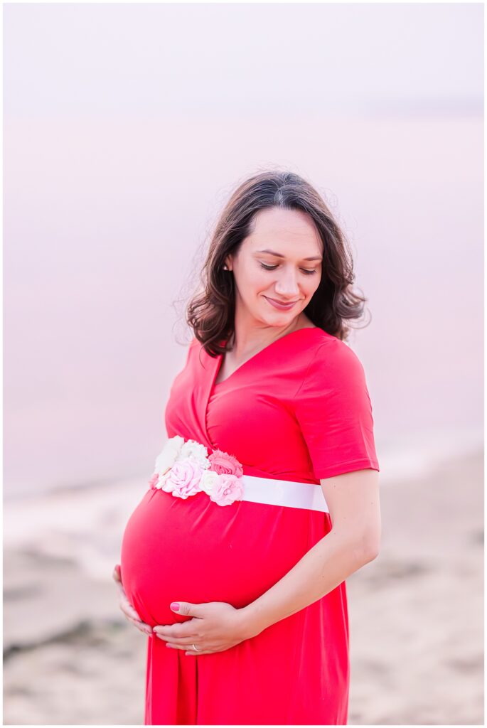 A close-up of the pregnant woman in a bright red dress, cradling her belly and looking down with a peaceful smile. The soft pink sky in the background adds a calm and beautiful touch to this maternity moment at Great Island Trail.