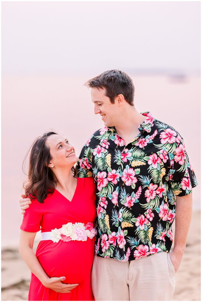 The couple smiles at each other on the beach, with the mother-to-be in a vibrant red dress and the man wearing a floral shirt. Their joy and connection create a warm and lively image perfect for Cape Cod maternity photography.