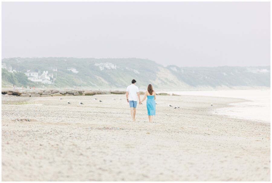 A couple walks hand-in-hand on a sandy beach in Sandwich, MA, with distant houses and hills in the background.