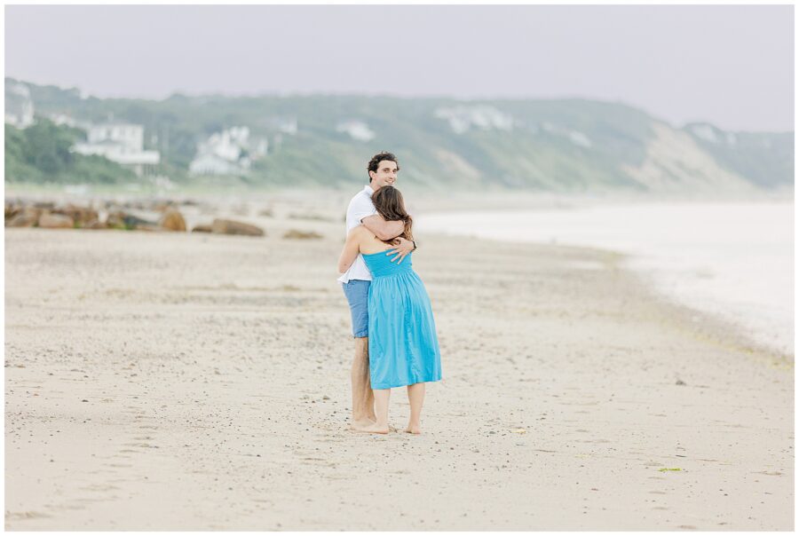 The couple embraces on the beach in Sandwich, MA, smiling with greenery and houses in the background.