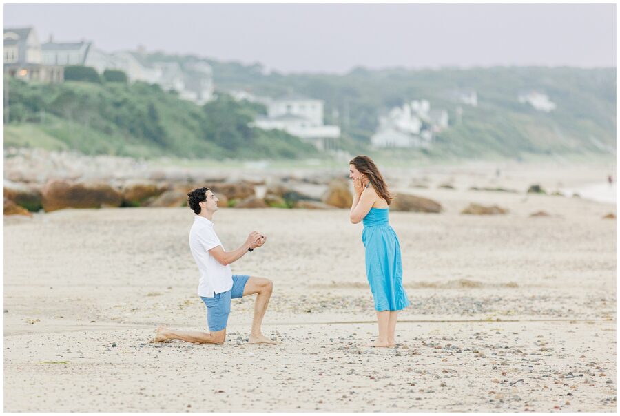 A man kneels and proposes to a surprised woman on the beach at Cape Cod, surrounded by rocks and distant houses.