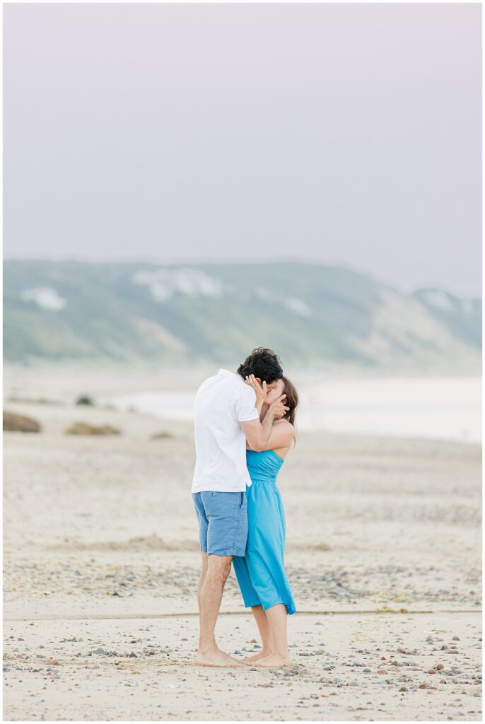 The couple kisses on the beach after the proposal, with hills in the background.