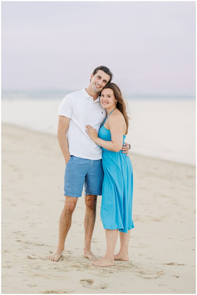 The couple poses, smiling and embracing, standing barefoot on the sandy beach.