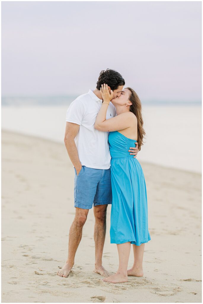 The couple kisses on the beach at Cape Cod, with the woman holding his face as they embrace.