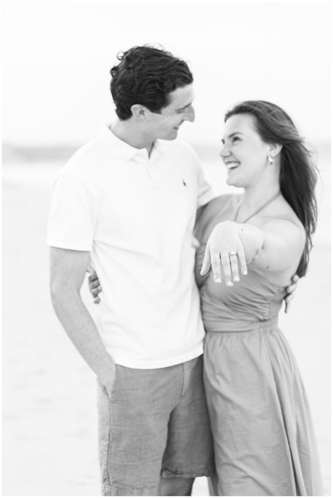 The woman displays her engagement ring, smiling at her partner on a Cape Cod beach.