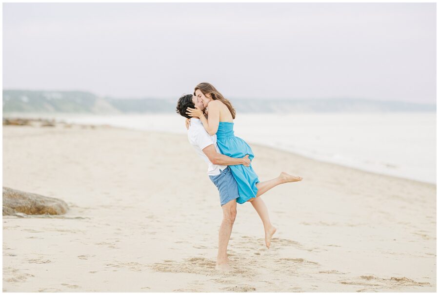 The man lifts the woman in an embrace on the Sandwich, MA beach as they kiss.