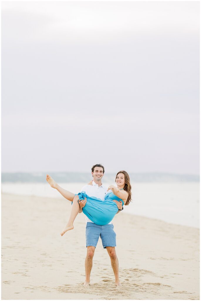 The man carries the woman in his arms on the beach at Sandwich, MA, both smiling with soft waves in the distance.