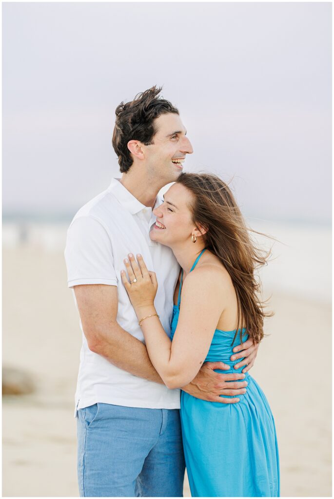 A couple smiles while embracing on the beach in Sandwich, MA, the woman wearing an engagement ring.