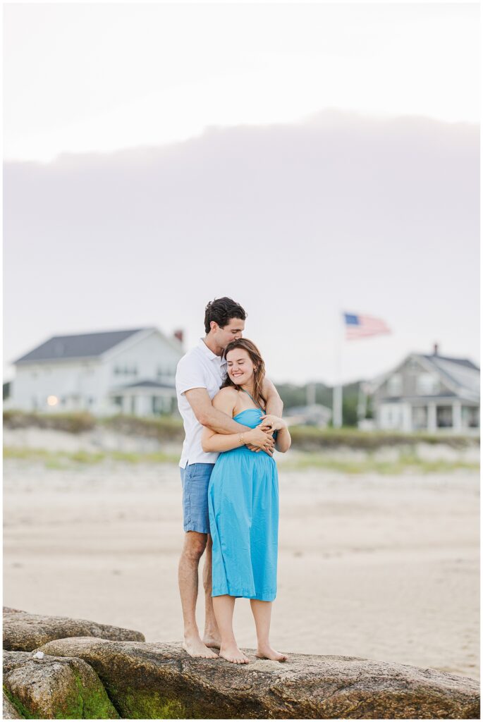 The couple stands on rocks by the beach in Sandwich, MA, hugging with houses and an American flag in the background.