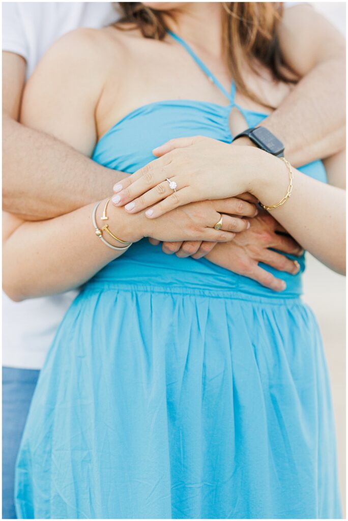 Close-up of the couple’s hands, showing the engagement ring and jewelry, with the woman in a blue dress.