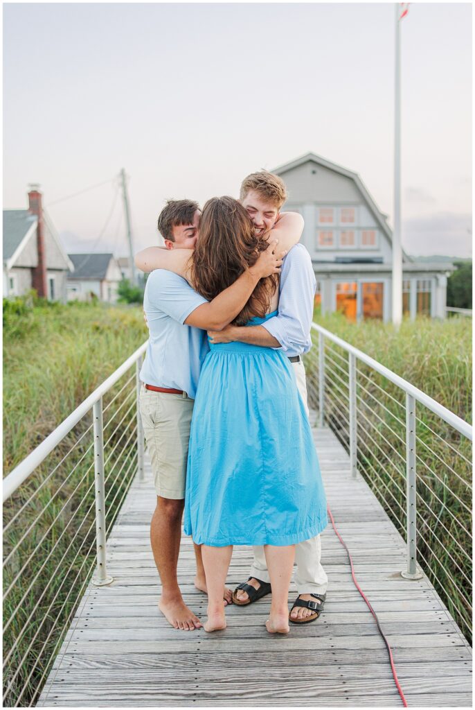 The woman hugs two people on a wooden walkway near beach houses in Sandwich, MA.