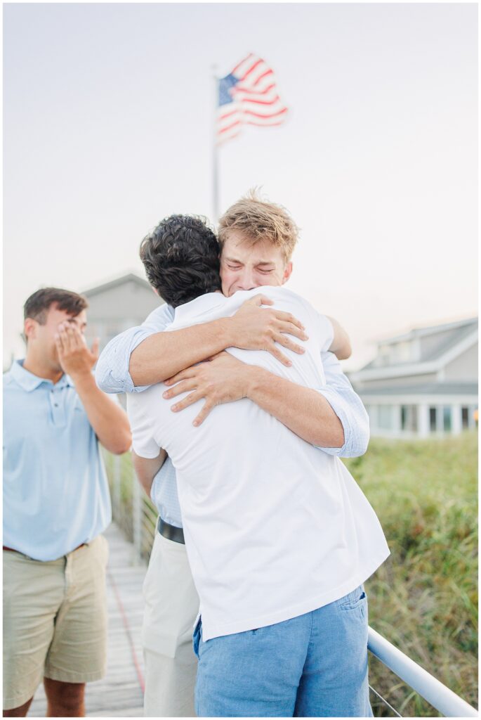 Two men embrace emotionally on a boardwalk in Sandwich, MA, with an American flag in the background.