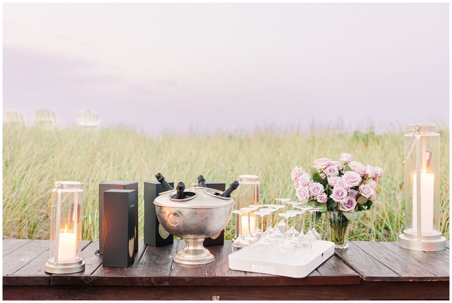 A table set with champagne, flowers, and candles for a celebration on a grassy beach in Sandwich, MA.