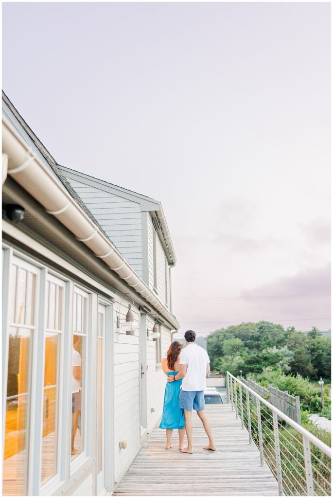 The couple walks arm-in-arm on a wooden deck in Sandwich, MA, with trees and greenery in the distance.