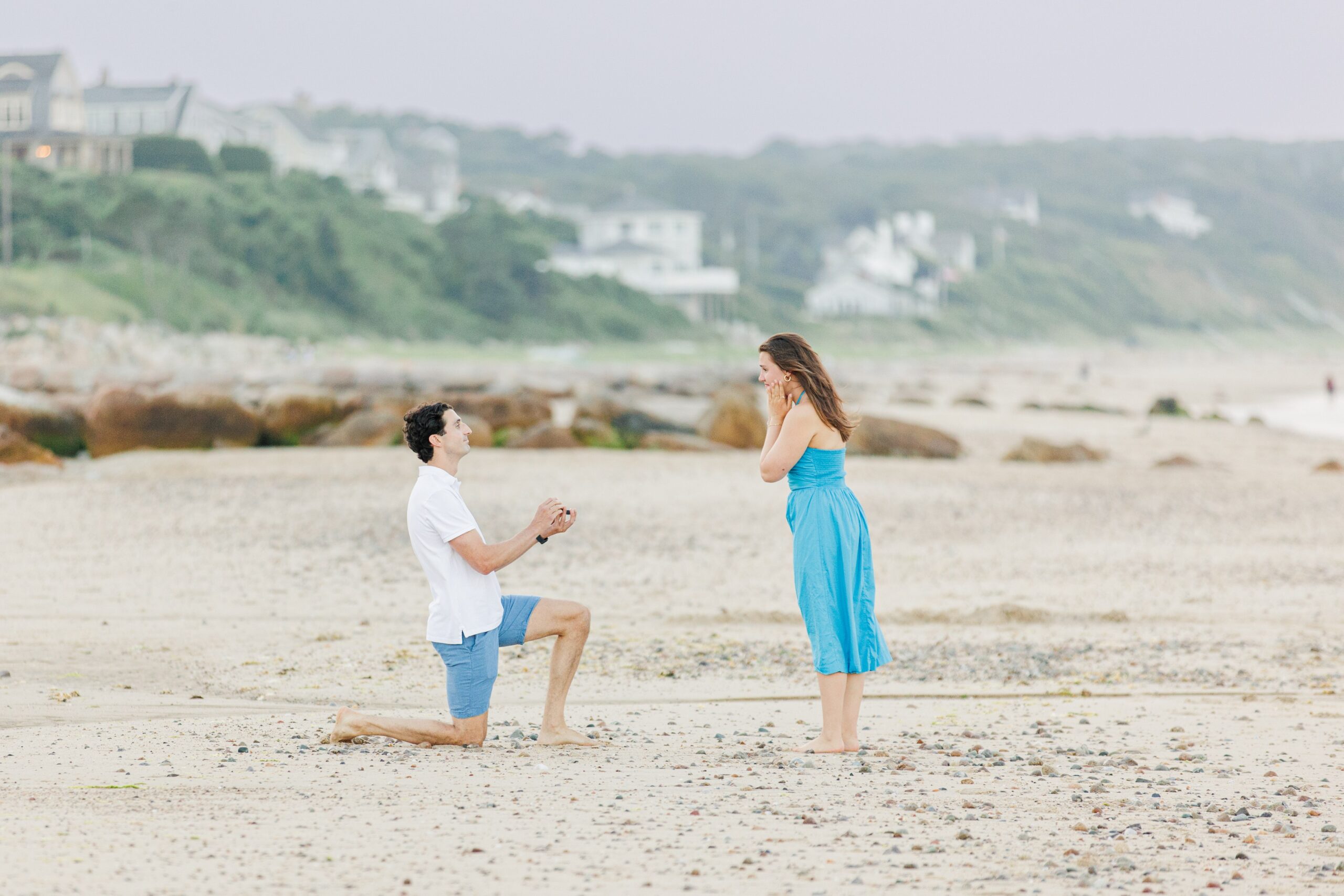 The man proposes on one knee on a sandy beach in Sandwich, MA, as the woman reacts in surprise.