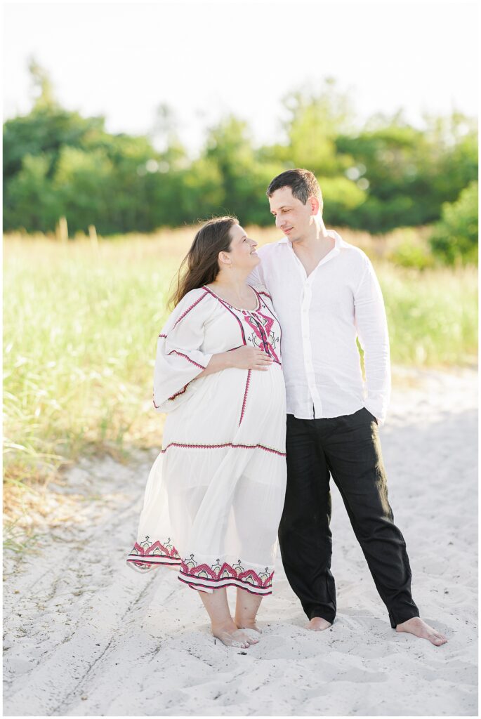 A pregnant woman and her partner stand on a sandy path with tall grass, gazing at each other. The woman wears a flowing, embroidered white dress, and the man wears a white shirt and black pants.