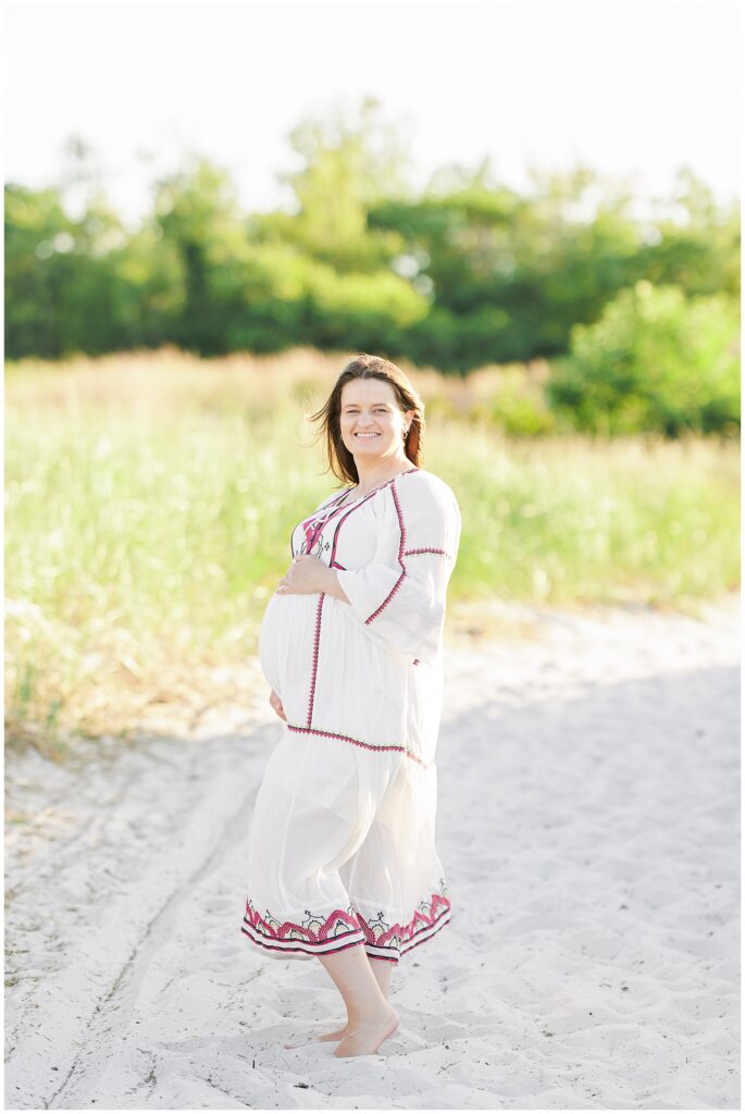 A pregnant woman smiles while standing barefoot on the beach, holding her belly, with tall grass and greenery behind her.