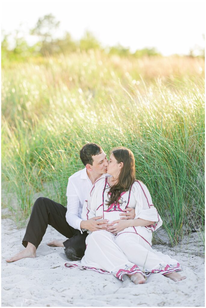 A couple sits on the sand, surrounded by grass, sharing a kiss. The woman is cradling her belly, and both are barefoot.