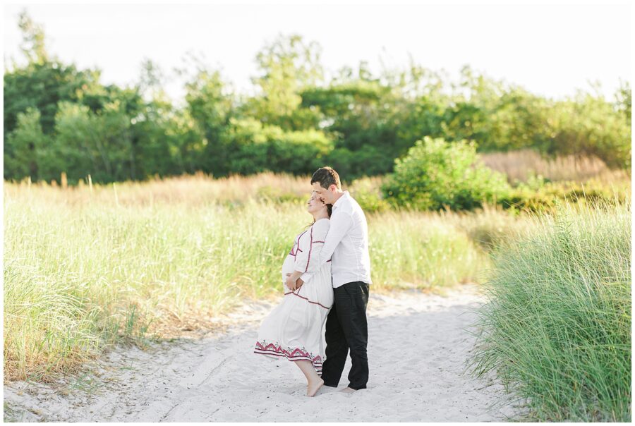 A couple stands together on a beach path surrounded by tall grass, the man hugging the woman from behind.