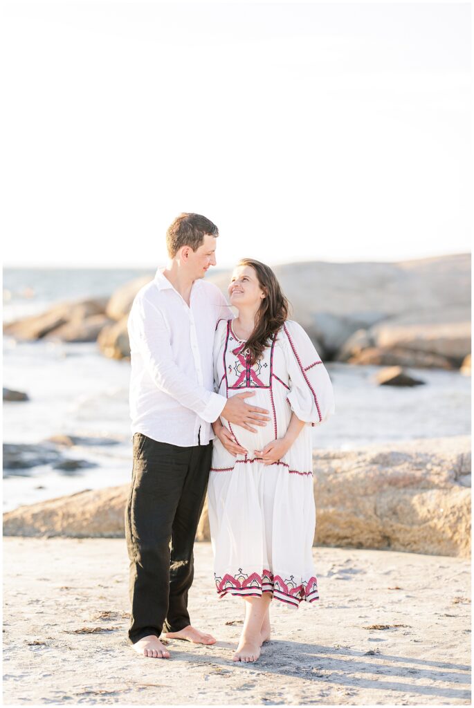 A couple stands on the beach with large rocks in the background, the man resting his hands on the woman’s pregnant belly as they look at each other.