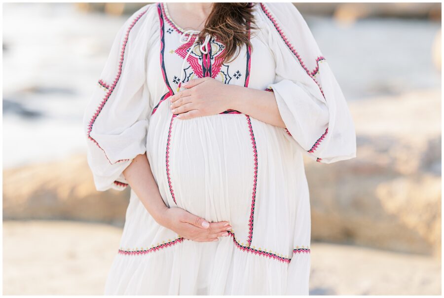 A close-up of the pregnant woman’s hands cradling her belly, focusing on the embroidered details of her dress.