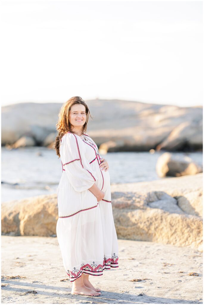 A pregnant woman in a white dress stands barefoot on a rocky beach with the ocean in the background.