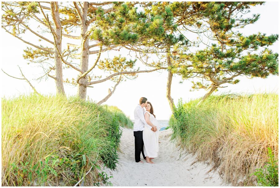 A couple standing on a sandy path surrounded by grass, with the man embracing the pregnant woman from behind.