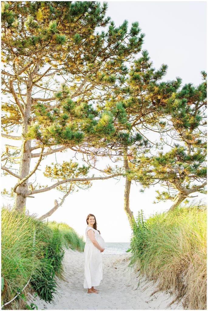 A pregnant woman in a white dress stands on a sandy beach path framed by two large trees.