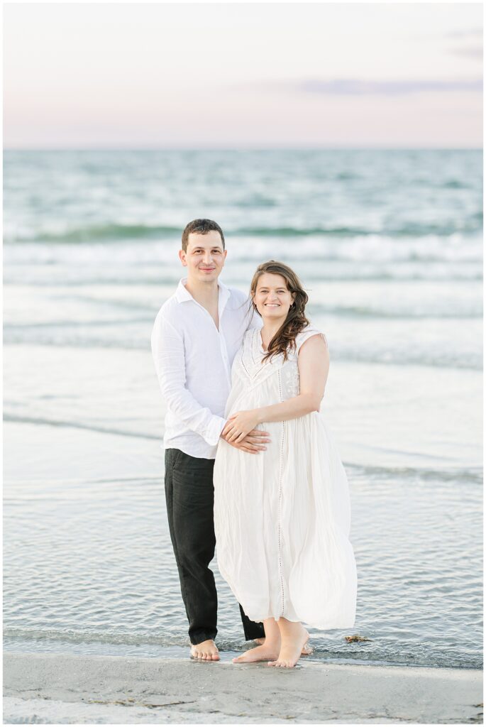 The couple stands together in shallow water, smiling at the camera, with the ocean behind them.