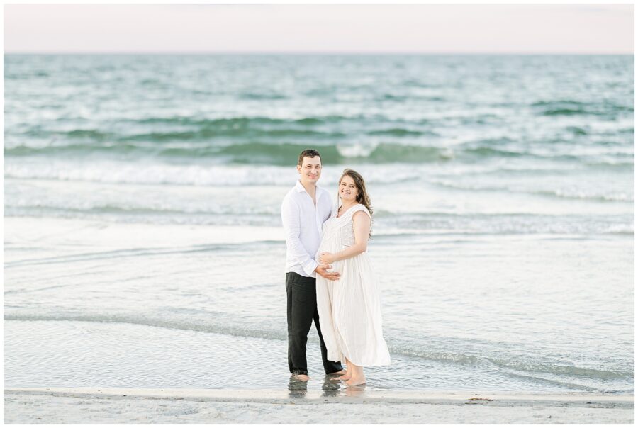 The couple stands together in shallow water, smiling at the camera, with the ocean behind them.