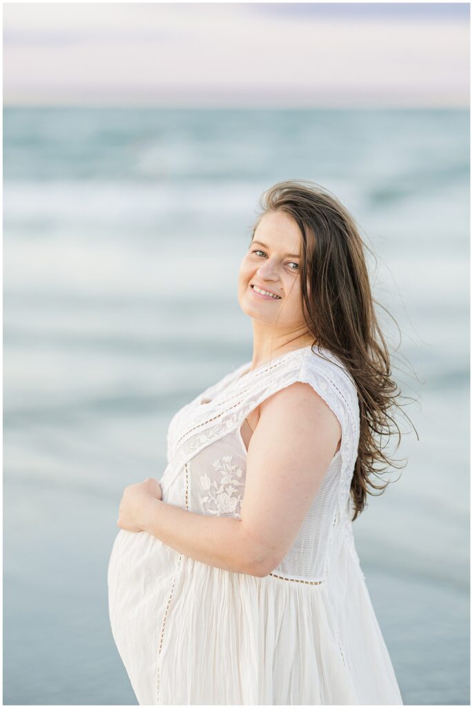 The woman smiles as she stands on the beach, holding her belly, with ocean waves in the background.