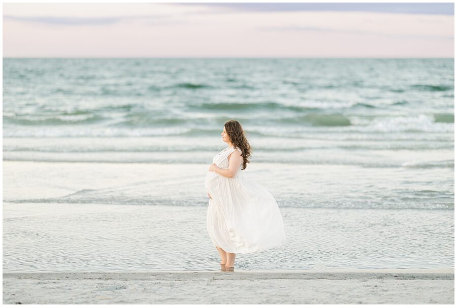 The woman stands alone at the water’s edge, holding her belly and gazing out at the ocean.