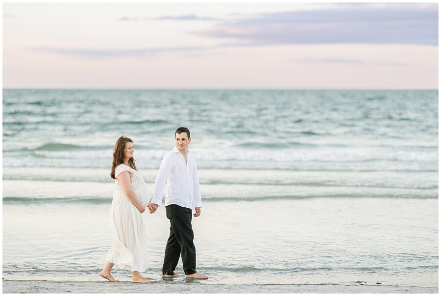 The couple holds hands and walks along the shoreline, the woman’s white dress flowing as they stroll barefoot on the wet sand.