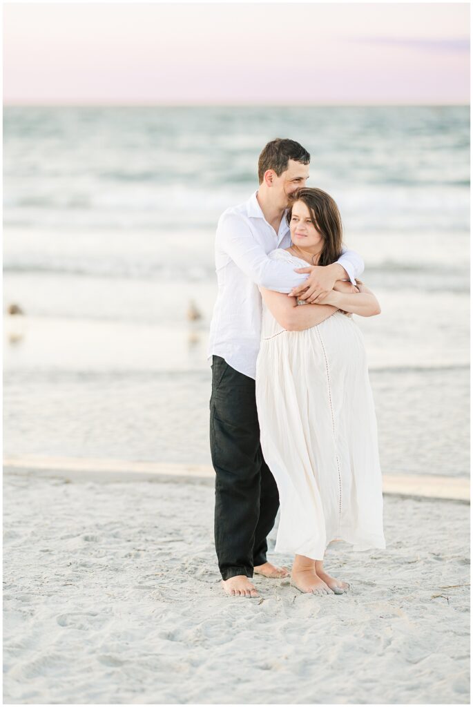 The man embraces the pregnant woman from behind as they stand barefoot on the beach, looking off into the distance.