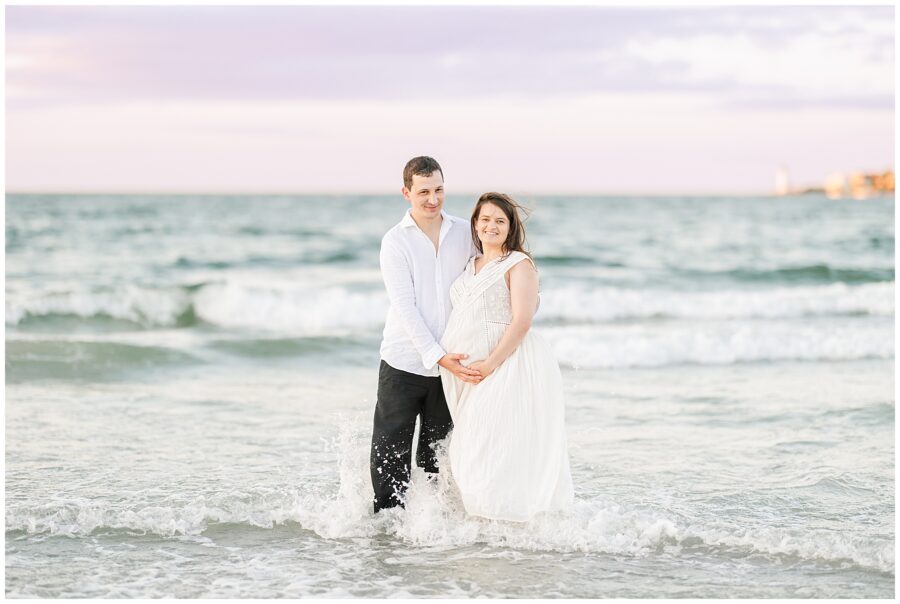 The couple stands together in the ocean, with small waves crashing at their feet. The man holds the woman’s belly while they smile at the camera.