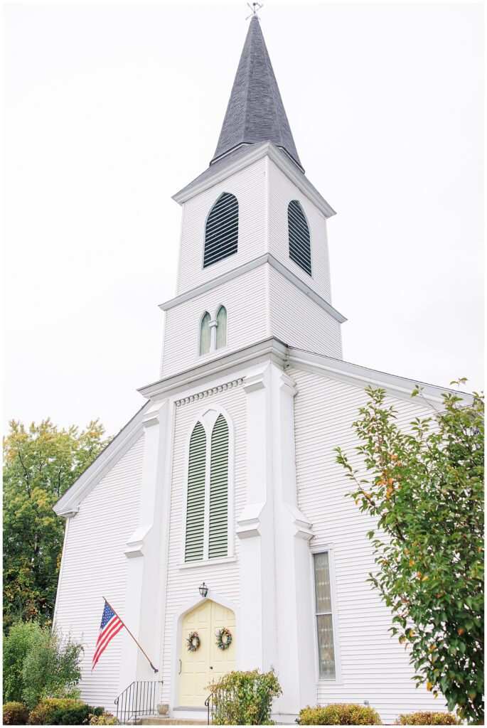 A tall white church building with a steeple and arched windows. The entrance has a double door with wreaths, and an American flag is mounted on the left side.