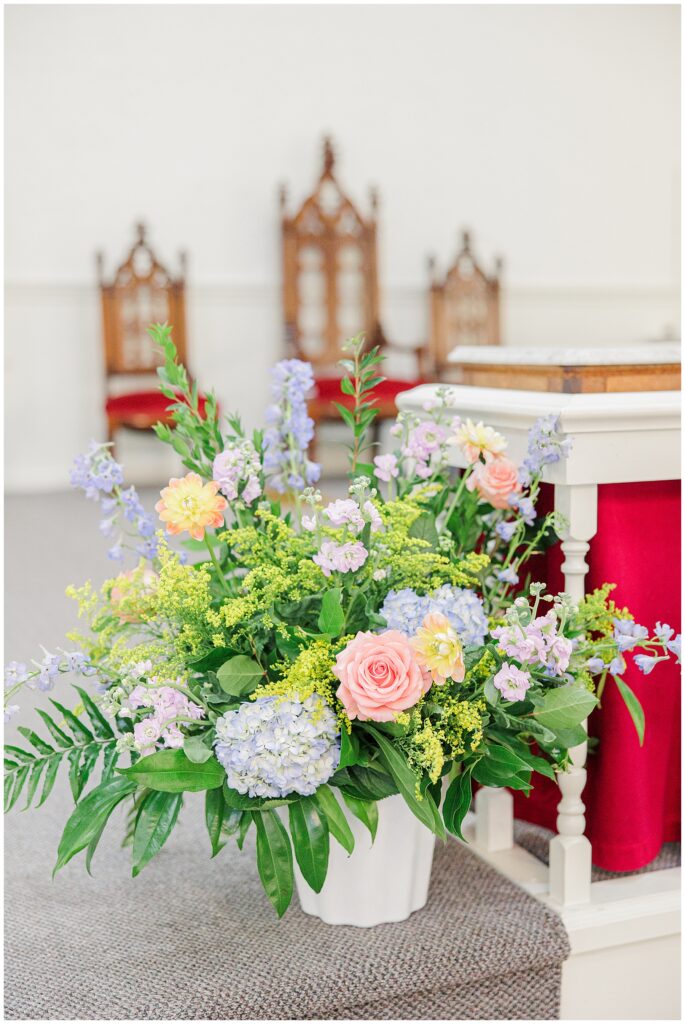 A large floral arrangement in a white vase, featuring roses, hydrangeas, and other flowers in pastel colors, placed near a pulpit.