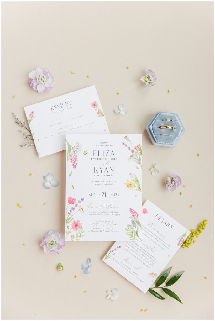 A flat lay of wedding invitations for Eliza Katherine Fisher and Ryan Scott Ardine, surrounded by flowers and wedding rings.