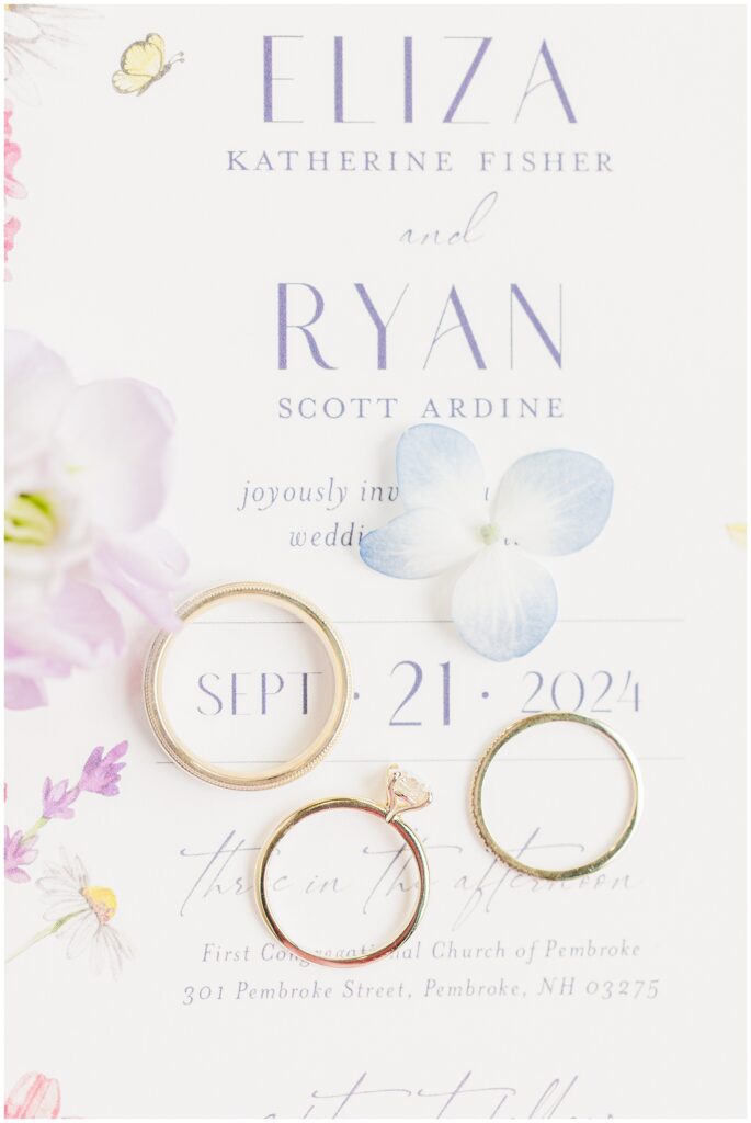 A flat lay of three gold rings (engagement and wedding bands) arranged on the wedding invitation.