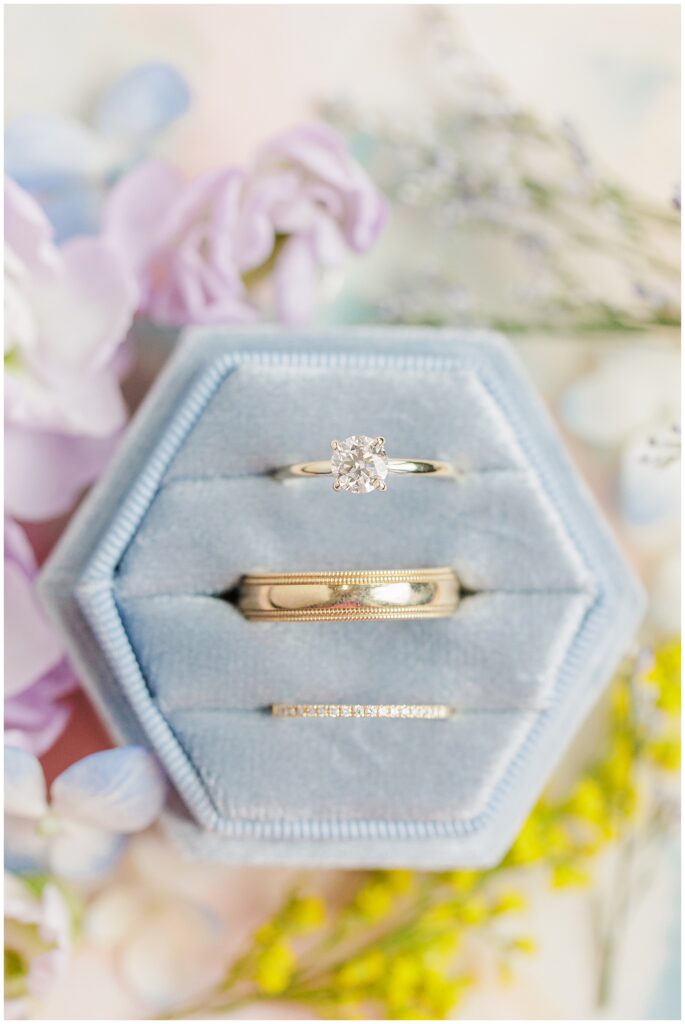 A close-up of the engagement ring, wedding bands, and purple flowers placed in a light blue ring box.